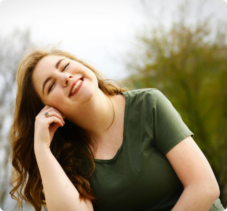 Young woman in a green shirt smiling with her head tilted back, outdoor background with blurred greenery, enjoying a break during her weight management routine.