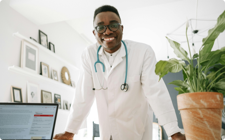 A cheerful male doctor in a white coat with a stethoscope around his neck, leaning over a desk with a computer in a bright office.