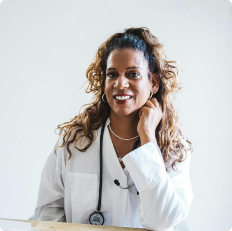 A smiling woman in a white lab coat with a stethoscope around her neck, standing in front of a white background.